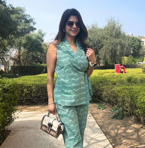 Woman standing outdoors in a garden, wearing a sleeveless green printed co-ord set (top and pants), holding a stylish handbag. The overall vibe is relaxed and fashionable.