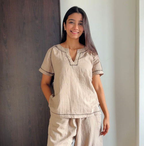 A woman is standing indoors against a wooden and white wall, smiling gently. She is wearing a comfortable beige co-ord set with a short-sleeve, V-neck top and matching relaxed-fit pants made of soft, crinkled fabric. Her hair is straight and left loose, and her pose reflects a casual, relaxed vibe.