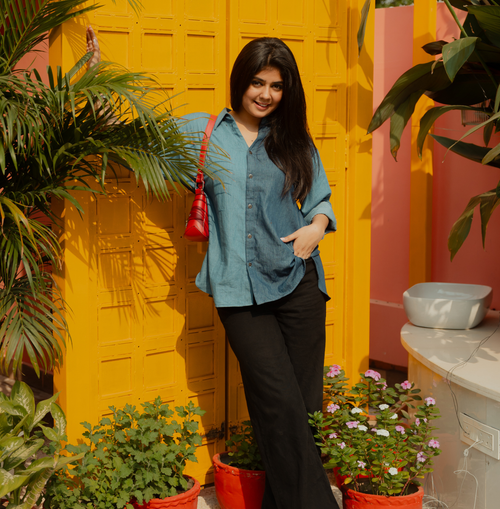 A woman is standing outdoors against a bright yellow textured door, wearing a loose blue button-down top with black pants and carrying a small red shoulder bag.
