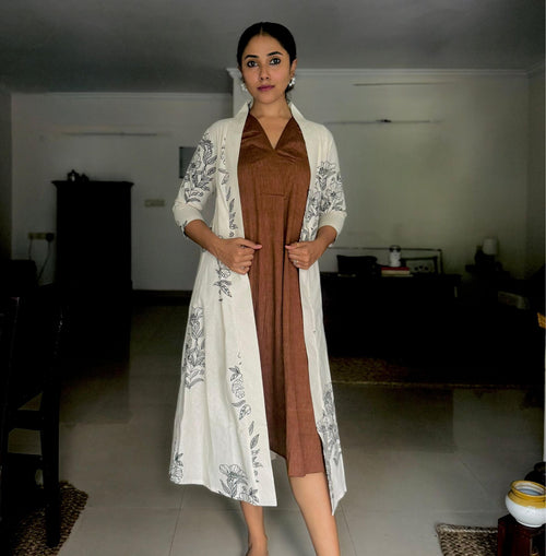 A woman stands indoors in a softly lit room, wearing a brown sleeveless dress with a pleated detail at the neckline. Over it, she has layered a long white shrug featuring black botanical prints. Her hair is neatly tied back, she wears statement earrings, and her pose is elegant and poised.